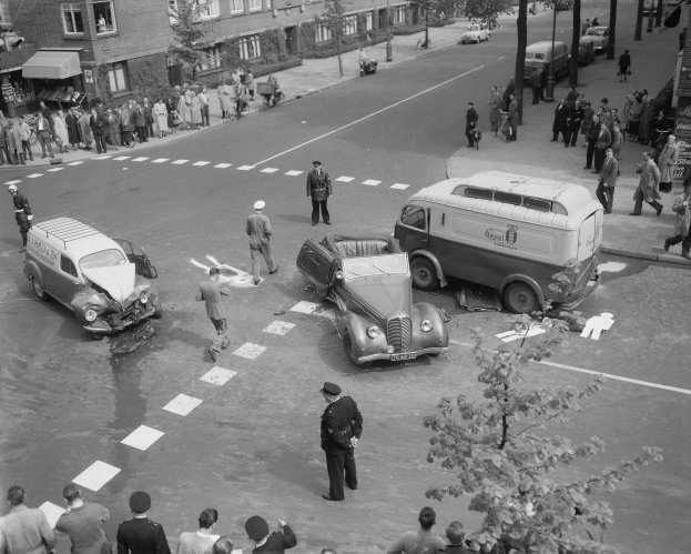 Schwarz-weiß-Szene eines Autounfalls an einer Straßenkreuzung mit mehreren Fahrzeugen, darunter ein Van, und einer Gruppe von Schaulustigen; Gebäude, Bäume, Strommasten und Texttafeln im Hintergrund.