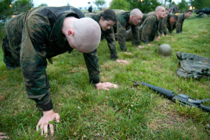 Eine Gruppe von Männern in Militäruniformen, die Liegestütze auf dem Gras machen, mit einer Waffe, einem Helm und anderen Gegenständen drumherum, mit Bäumen, Lichtern, Fahrzeugen und einem klaren blauen Himmel im Hintergrund.