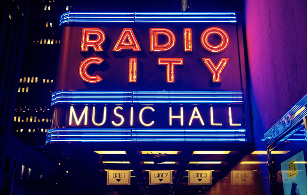 Außensicht des Radio City Music Hall in New York City bei Nacht, hell erleuchtet mit hohen Gebäuden im Hintergrund, mit einer 'Radio City'-Schrifttafel und zusätzlichen Schildern auf der rechten Seite.