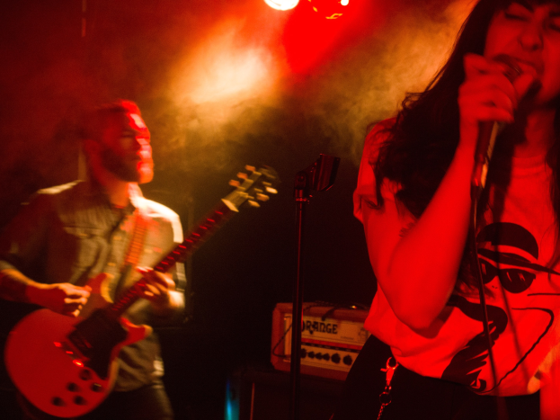Eine Frau singt in ein Mikrofon auf der Bühne, während ein Mann im Hintergrund Gitarre spielt.
