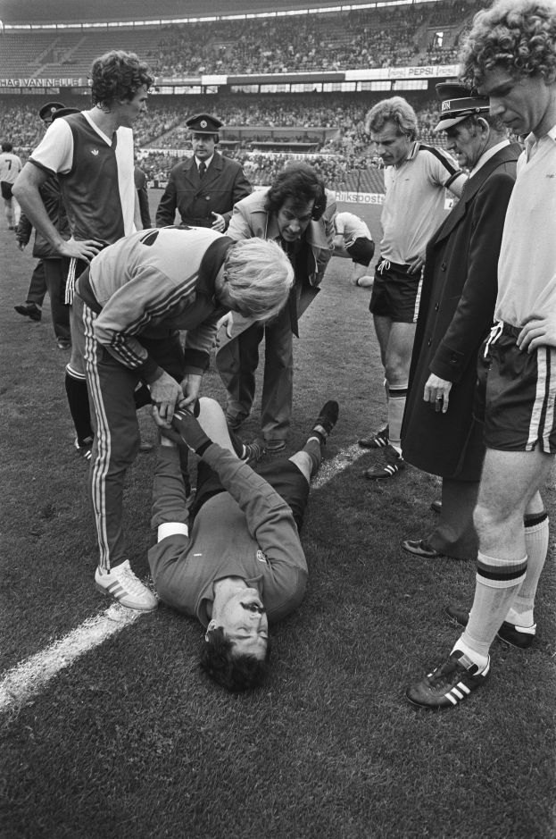 Schwarzes und weißes Bild eines Fußballspielers auf dem Boden umgeben von einer Gruppe von Männern, mit einer Menge im Stadionhintergrund.