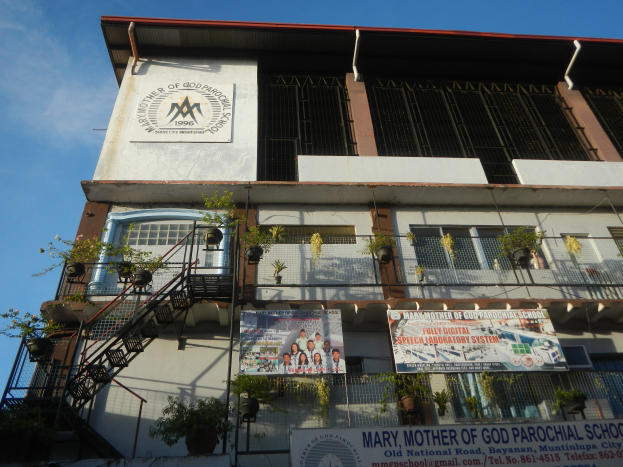 Das Gebäude der Mariä Himmelfahrt Grundschule mit Fenstern, Geländern, Treppen, Topfpflanzen, Bannern und einem bewölkten Himmel im Hintergrund.