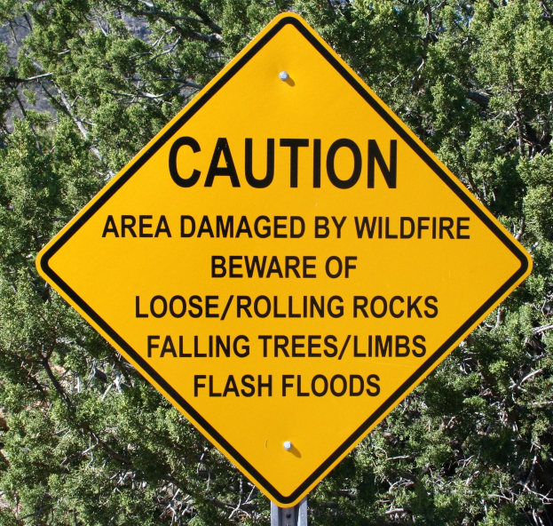Gelbes Warnschild an einem Pfahl im Vordergrund, das vor Wildfire-Schäden, lockeren Steinen, umstürzenden Bäumen und Überschwemmungen warnt, mit Bäumen im Hintergrund.