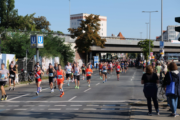 Gruppe von Menschen, die bei einem Marathon auf einer von Bäumen gesäumten Straße laufen, mit Fahrrädern, Schildern, einem Zaun, Gras, einer Brücke, Gebäuden und einem klaren blauen Himmel.