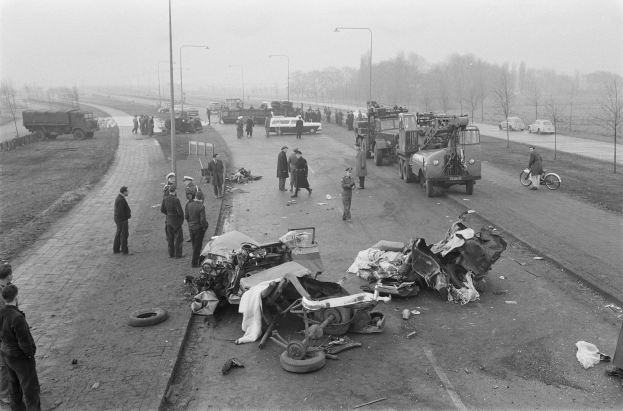 Schwarz-weißes Bild einer Autounfall-Szene am Straßenrand mit mehreren Fahrzeugen und einer Gruppe von Menschen drumherum, Lichtmasten, Bäume und Himmel im Hintergrund.