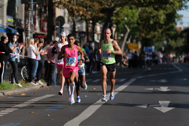 Gruppe von Läufern bei einem Marathon auf einer Stadtstraße mit Zuschauern auf der linken Seite, unscharfe Bäume, Gebäude, Pfosten, Schilder und ein Fahrrad im Hintergrund.