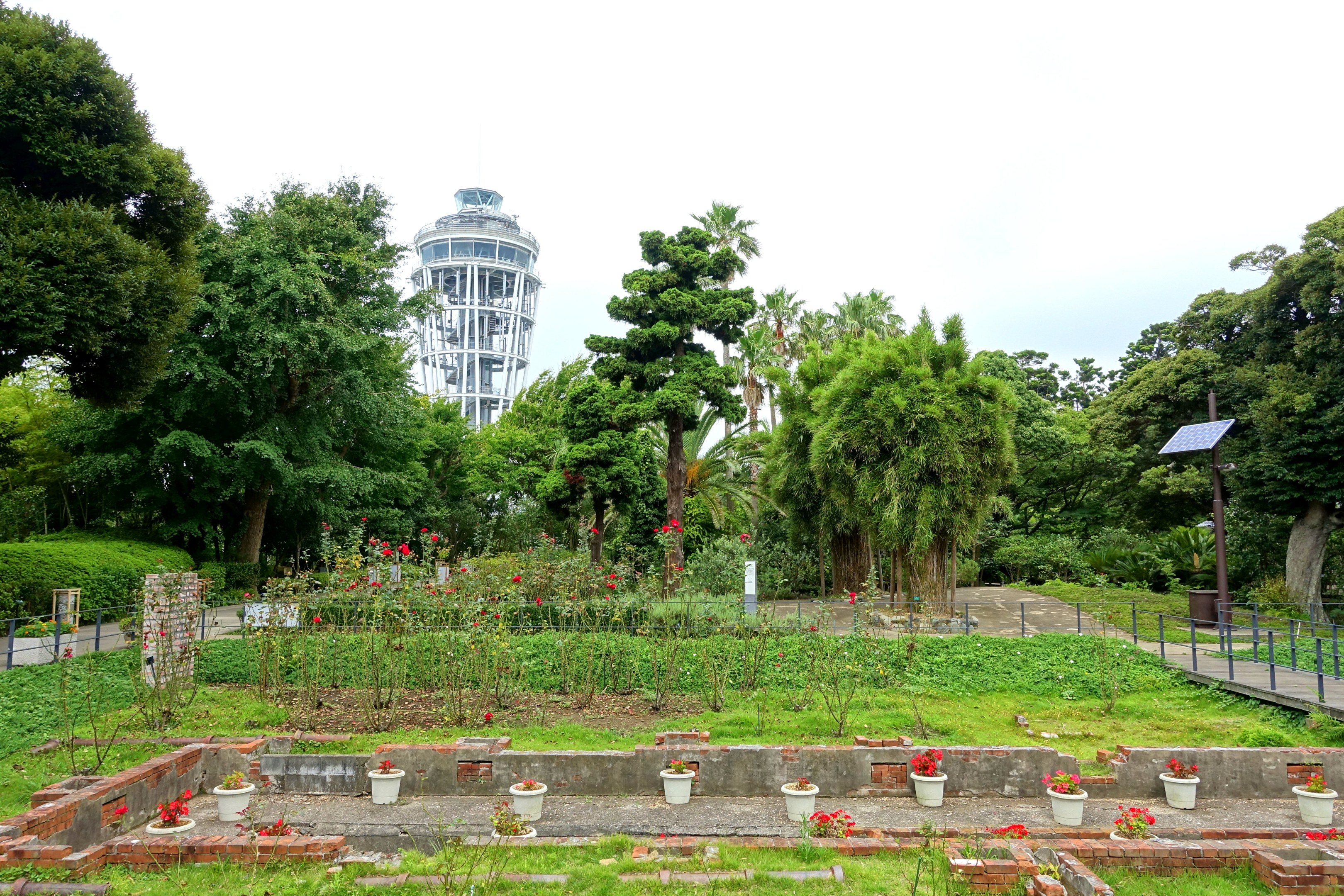 Ein Park mit einem Turm im Hintergrund, umgeben von grünem Gras, Pflanzen, Bäumen und verstreuten Blumentöpfen, mit einer Straße mit Geländern und einer an einem Pfahl befestigten Tafel unter einem sichtbaren Himmel.
