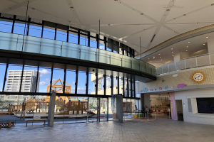 Innenansicht des Haupt входs zu einem neuen Kinderspielbereich mit großen Fenstern, elektrischen Lampen, einer Wanduhr, einem Fernseher, einem Anzeigebildschirm, hölzernen Gegenständen und einem Zaun, umgeben von Gebäuden, Bäumen und einem bewölkten Himmel.