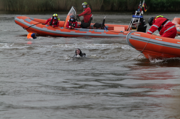 Eine Gruppe von Menschen in einem aufblasbaren Boot auf einem Fluss, mit zwei Personen im Wasser im Vordergrund und Vegetation im Hintergrund, alle tragen Schwimmwesten und Helme während einer Rettungsaktion.