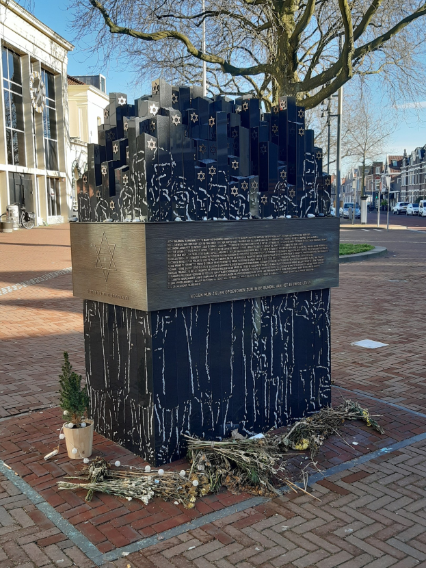 Denkmal in einem Stadtplatz gewidmet an Holocaust-Opfern, mit einem Baum davor, einer Topfpflanze, verstreuten trockenen Blättern, umgeben von Gebäuden, Fahrzeugen, Strommasten und Bäumen unter einem Himmel.