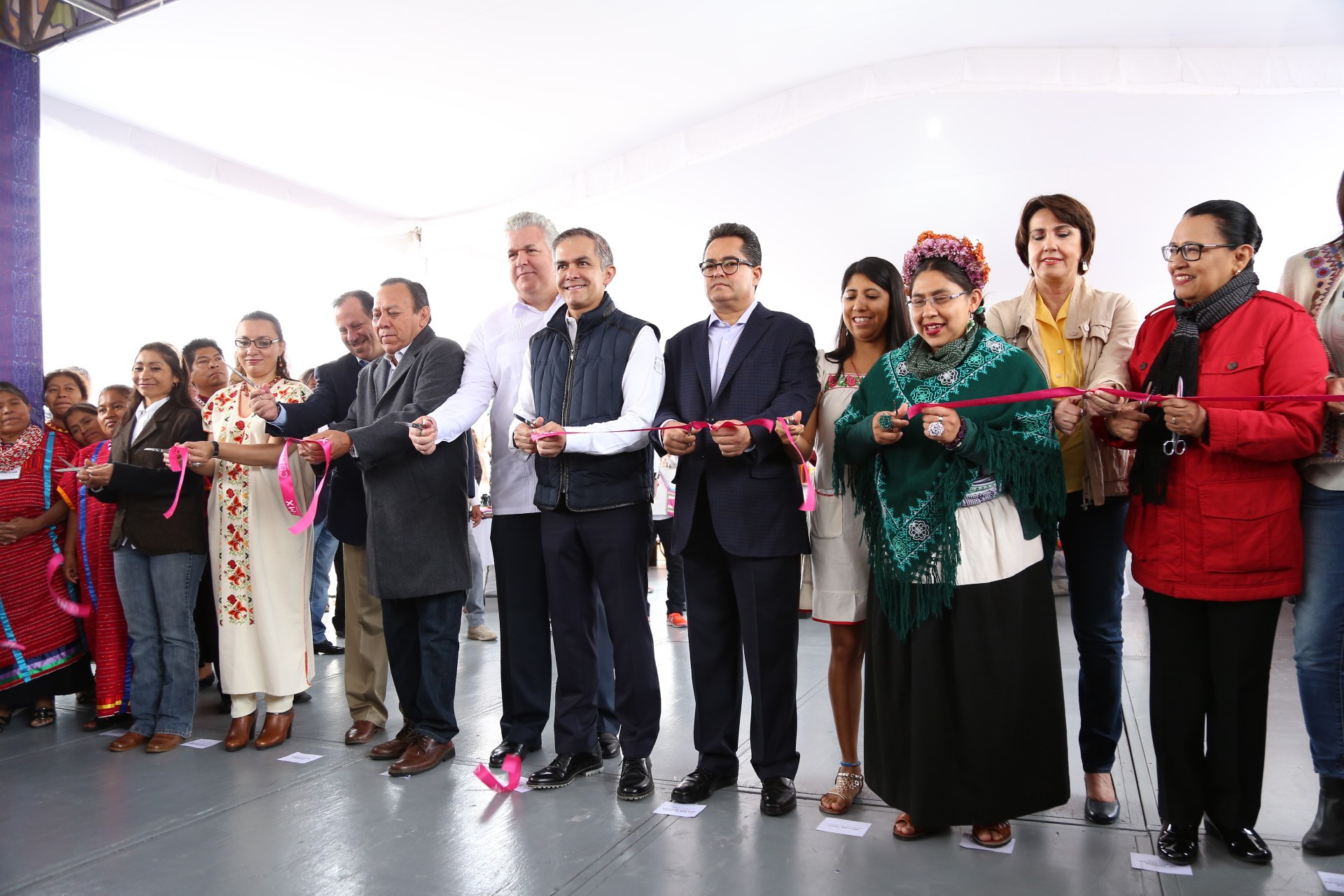 Eine Gruppe von Menschen, die strahlend eine Schere halten und vor einer Menge stehen, mit einer Säule links daneben, Papieren auf dem Boden und einem weißen Tuch im Hintergrund.