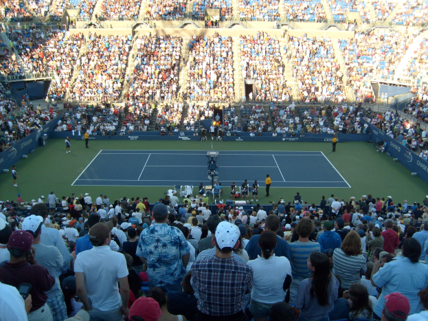 Große Zuschauermenge bei einem Tennisspiel in einem vollen Stadion mit Spielern auf dem Platz und Zuschauern in den Rängen.