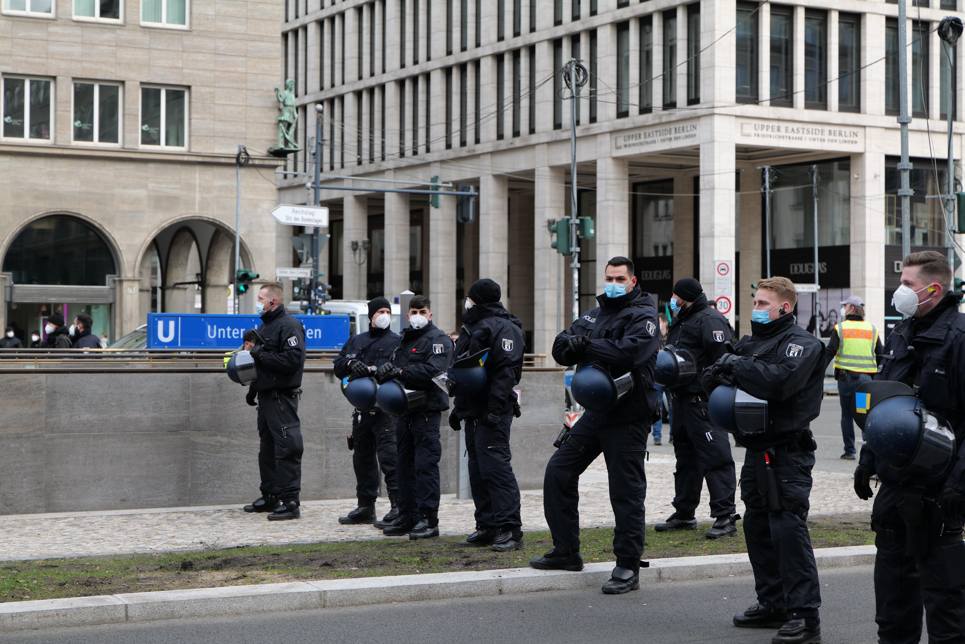 Eine Gruppe von Polizisten in schwarzen Uniformen und Masken steht vor einem Berliner Gebäude mit Glasfenstern und Säulen, einige halten Helme, mit Laternen, Verkehrsampeln, Texttafeln, einer Statue und anderen Menschen im Hintergrund auf einer grasbewachsenen Fläche.