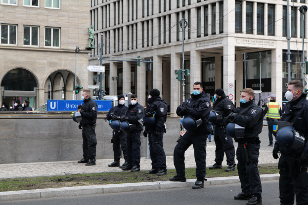 Eine Gruppe von Polizisten in schwarzen Uniformen und Masken steht vor einem Berliner Gebäude mit Glasfenstern und Säulen, einige halten Helme, mit Laternen, Verkehrsampeln, Texttafeln, einer Statue und anderen Menschen im Hintergrund auf einer grasbewachsenen Fläche.