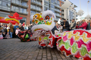 Ein lebendiges chinesisches Neujahrsfest in Amsterdam mit einer Löwen-Tanz-Show und einer Menge Schaulustiger, einige fotografieren das Ereignis, vor einem Gebäude und einem klaren blauen Himmel.