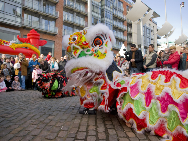 Ein lebendiges chinesisches Neujahrsfest in Amsterdam mit einer Löwen-Tanz-Show und einer Menge Schaulustiger, einige fotografieren das Ereignis, vor einem Gebäude und einem klaren blauen Himmel.