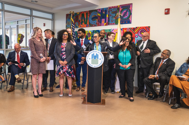 Gruppe von Menschen vor einem Podium mit Mikrofon und einer Tafel mit der Aufschrift "Sancho Stadtverordneter tritt dem Stadtrat bei", mit sitzenden und stehenden Teilnehmern, Flaggen, Feuerlöscher und Deckenleuchten im Hintergrund.