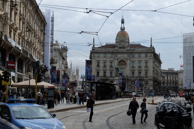 Eine belebte Stadtstraße mit einem geparkten Polizeiwagen, Fußgängern mit Taschen, fahrenden Fahrzeugen, Gebäuden mit Fenstern und Bannern, Laternen, Verkehrsampeln und einem bewölkten Himmel.