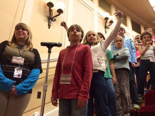 Eine Gruppe von Kindern singt an einem Mikrofon mit einem Erwachsenen daneben, in einem Raum mit Stühlen und beleuchteten Wandpaneelen im Hintergrund.