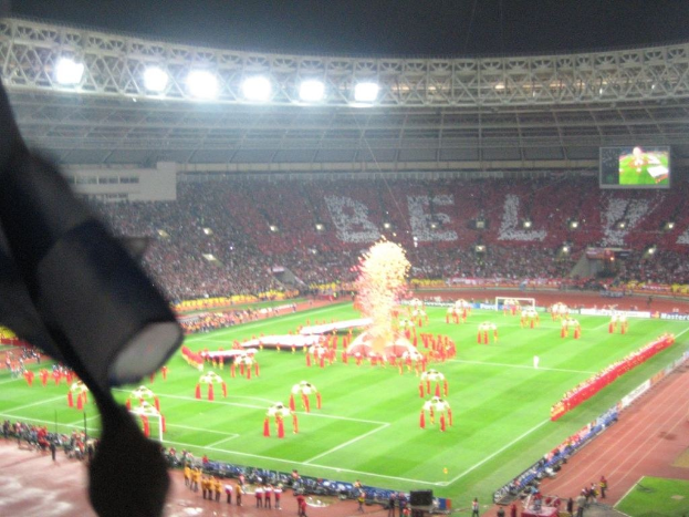 Ein Stadion mit Flutlicht, Eisengittern, Zuschauern, stehenden Personen, Werbetafeln, Anzeigetafeln, Überwachungskamera, Flaggen und Dekorationen.