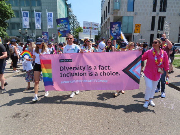 Eine Gruppe von Menschen marschiert auf einer Straße, hält ein Banner mit der Aufschrift "Diversität ist eine Tatsache, Inklusion ist eine Wahl" und trägt teilweise Mützen und Schutzbrillen, während andere zusätzliche Banner halten.