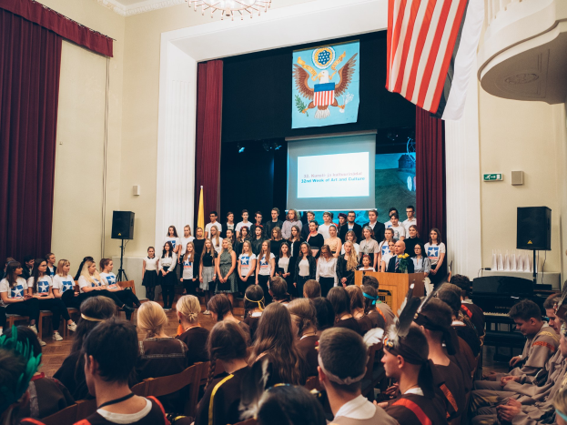 Eine Gruppe von Menschen steht vor einem Publikum mit Mikrofon auf einem Podium, flankiert von Fahnen und Lautsprechern, während einer kulturellen Veranstaltung mit einem Bildschirm im Hintergrund, der Text und Bilder anzeigt.