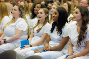 Eine Gruppe von Frauen in weißen Kleidern sitzt in einem Hörsaal, jede hält ein blaues Objekt in der Hand, mit einem unscharfen Hintergrund, der die Frauen betont, die scheinbar Teil eines Pflegeprogramms sind.