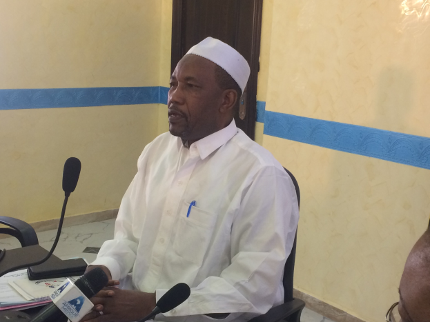 Ein Mann in einem weißen Kleid und Hut sitzt an einem Tisch mit Mikrofonen, spricht, während Papiere und Gegenstände auf dem Tisch liegen, mit einem anderen Mann mit Brille rechts von ihm in einem Raum mit einer Wand und einer Tür.