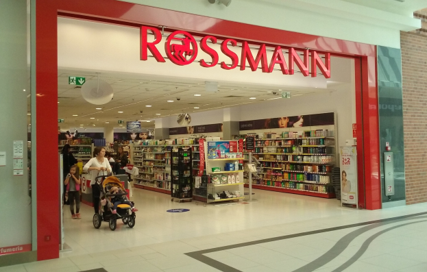 Ein Rossmann-Laden in einem Einkaufszentrum mit Menschen auf dem Boden, einer mit einem Kinderwagen, und Regalen mit verschiedenen Artikeln unter Deckenleuchten und Textschildern.