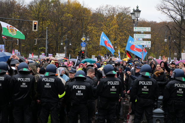 Eine große Gruppe von Polizisten steht vor einer Menge mit Schirmen, Fahnen und Schildern, mit Laternenmasten, Verkehrsampeln, Bäumen und einem klaren Himmel im Hintergrund.