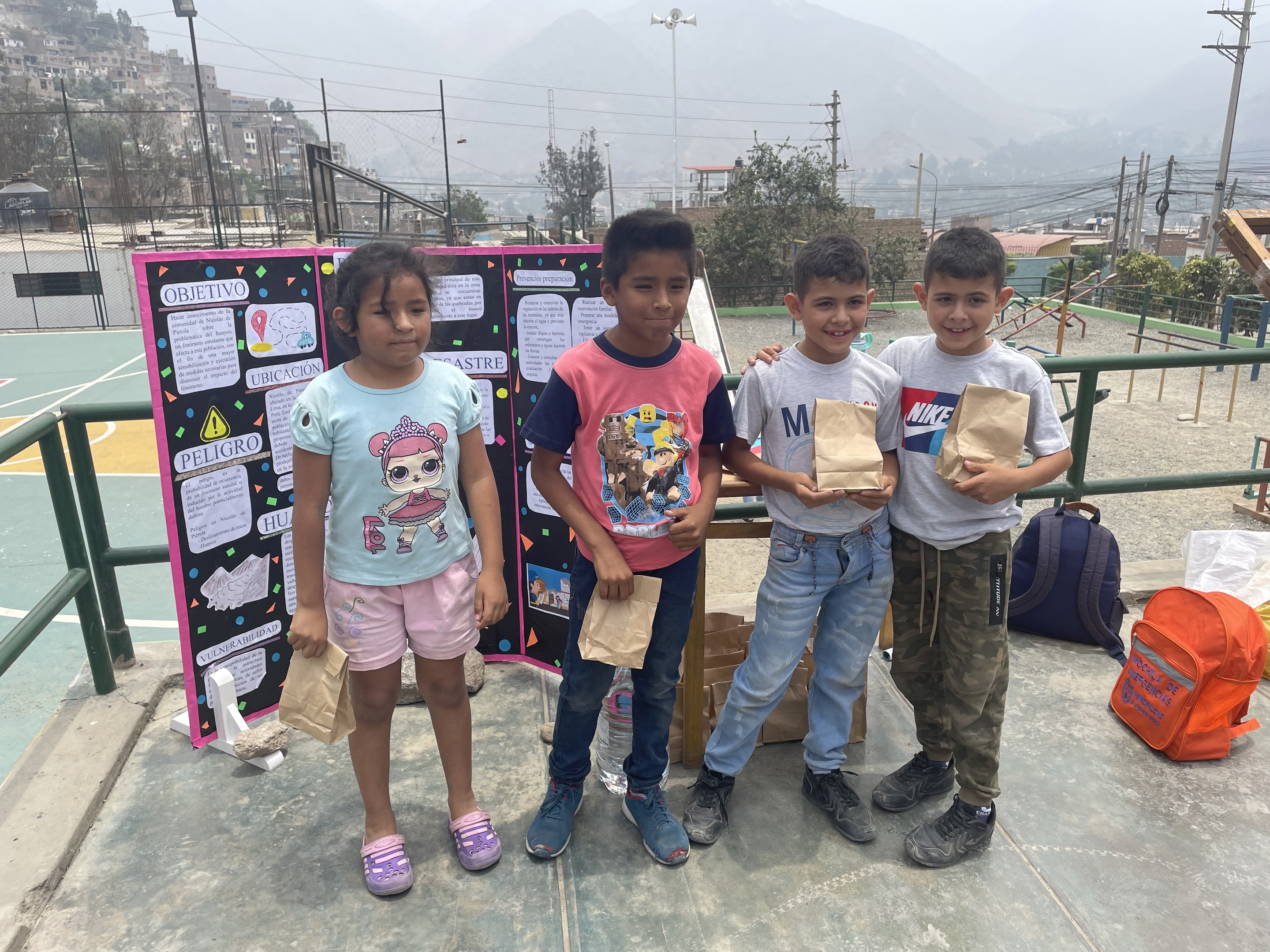 Gruppe von Kindern auf einem Basketballfeld mit Papiertüten in den Händen, Gegenstände auf dem Boden und eine Tafel mit Text links daneben, vor einem Hintergrund aus Zaun, Pfosten, Straßenlaternen, Bäumen, Gebäuden, Hügeln und Himmel.