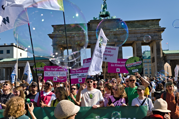 Menschenmenge vor dem Brandenburger Tor in Berlin, viele mit Mützen und Schutzbrillen, einige mit Fahnen und Transparenten mit Text, im Hintergrund Gebäude, ein von einer Statue gekrönter Bogen und ein mit Seifenblasen gefüllter Himmel.