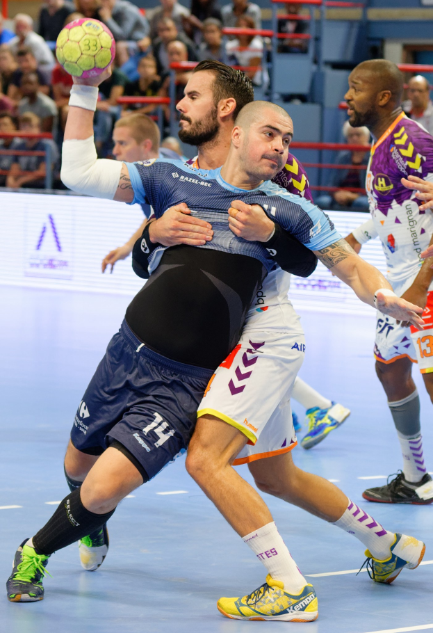 Männer spielen Handball in einem Stadion, einer hält den Ball, mit Zuschauern im Hintergrund.