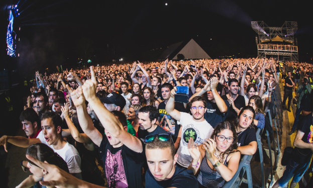 Menschenmenge auf einem Musikfestival mit erhobenen Händen, sitzend und stehend, neben einem Metallzaun, mit Gebäuden und Lichtern im Hintergrund unter einem dunklen Himmel.