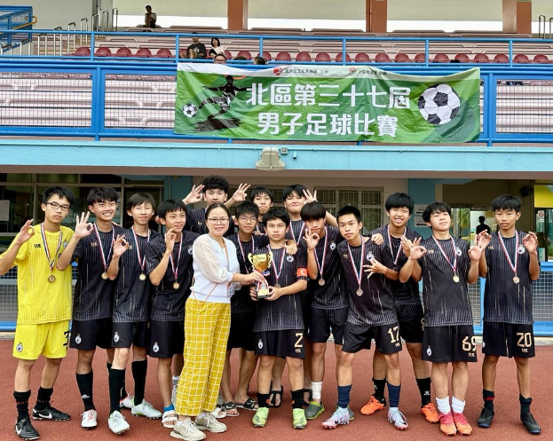 Gruppe junger Männer in Fußballtrikots auf einem Feld mit einem Pokal und Medaillen, im Hintergrund eine 'Yokohama U-16 Jungenfußballmannschaft'-Tafel und Zuschauer in der Nähe sitzend.