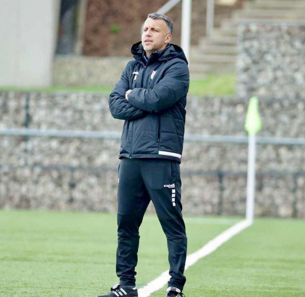 Ein Fußballtrainer auf einem Feld mit verschränkten Armen, der eine schwarze Jacke und Hose trägt, vor einem Zaun, einer Treppe und einer Wand.