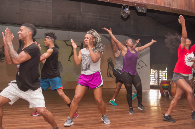 Gruppe von Menschen, die in einem Studio mit einer bemalten Wand, Fenster und Deckenbeleuchtung tanzen, wahrscheinlich während einer Zumba-Stunde.