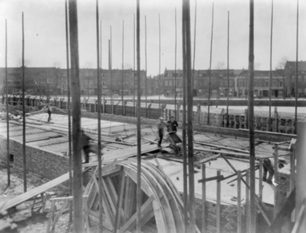 Schwarzes und weißes Foto einer Baustelle mit Arbeitern, Holzpfählen und einem Rad im Vordergrund, Gebäuden, Bäumen und einem klaren Himmel im Hintergrund.