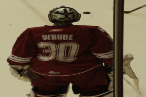 Ein Hockey-Spieler in Helm, Handschuhen und nummerierter Weste steht auf dem Eis vor einem Tor mit einem Pfosten rechts daneben.