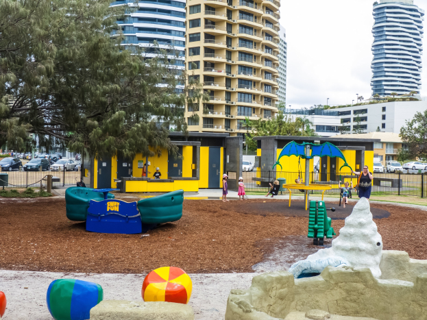 Spielplatz in städtischer Umgebung mit Kindern auf Geräten, umgeben von Bäumen und Gebäuden, mit Fahrzeugen auf der Straße und Himmel darüber.