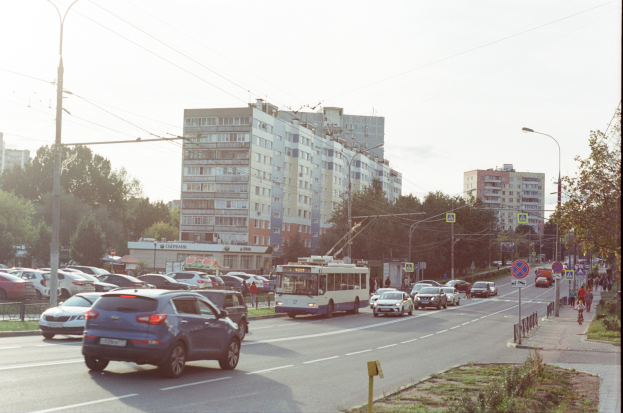 Eine belebte Stadtstraße mit Autos, Bussen, Straßenlaternen, Schildern, Laternenmasten, Strommasten mit Drähten, Bäumen, Gebäuden mit Fenstern und einem klaren blauen Himmel.