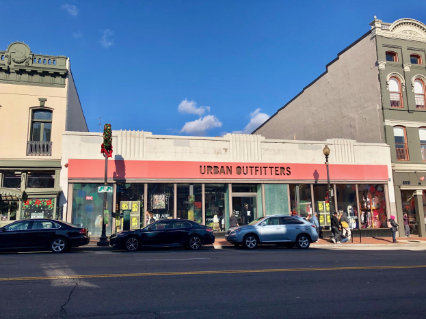 Eine belebte Stadtstraße mit parkenden Autos, Gebäuden mit Fenstern und einer "Urban Outfitters"-Reklame, Laternenpfählen, Fußgängern auf dem Gehweg und einer bewölkten Himmel im Hintergrund.