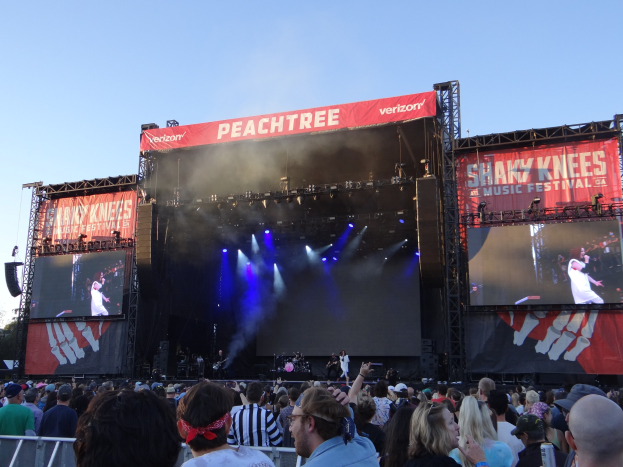 Eine große Menschenmenge vor einer hell erleuchteten Bühne beim Shaky Knees Music Festival, wo Musiker hinter einem Geländer unter einem klaren blauen Himmel spielen.