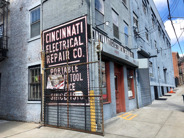 Außenansicht des Cincinnati Electrical Repair Co.-Gebäudes mit einem Schild, einem Metallzaun, einem Müllcontainer, Pfählen, Drähten, Bäumen und einem bewölkten Himmel.