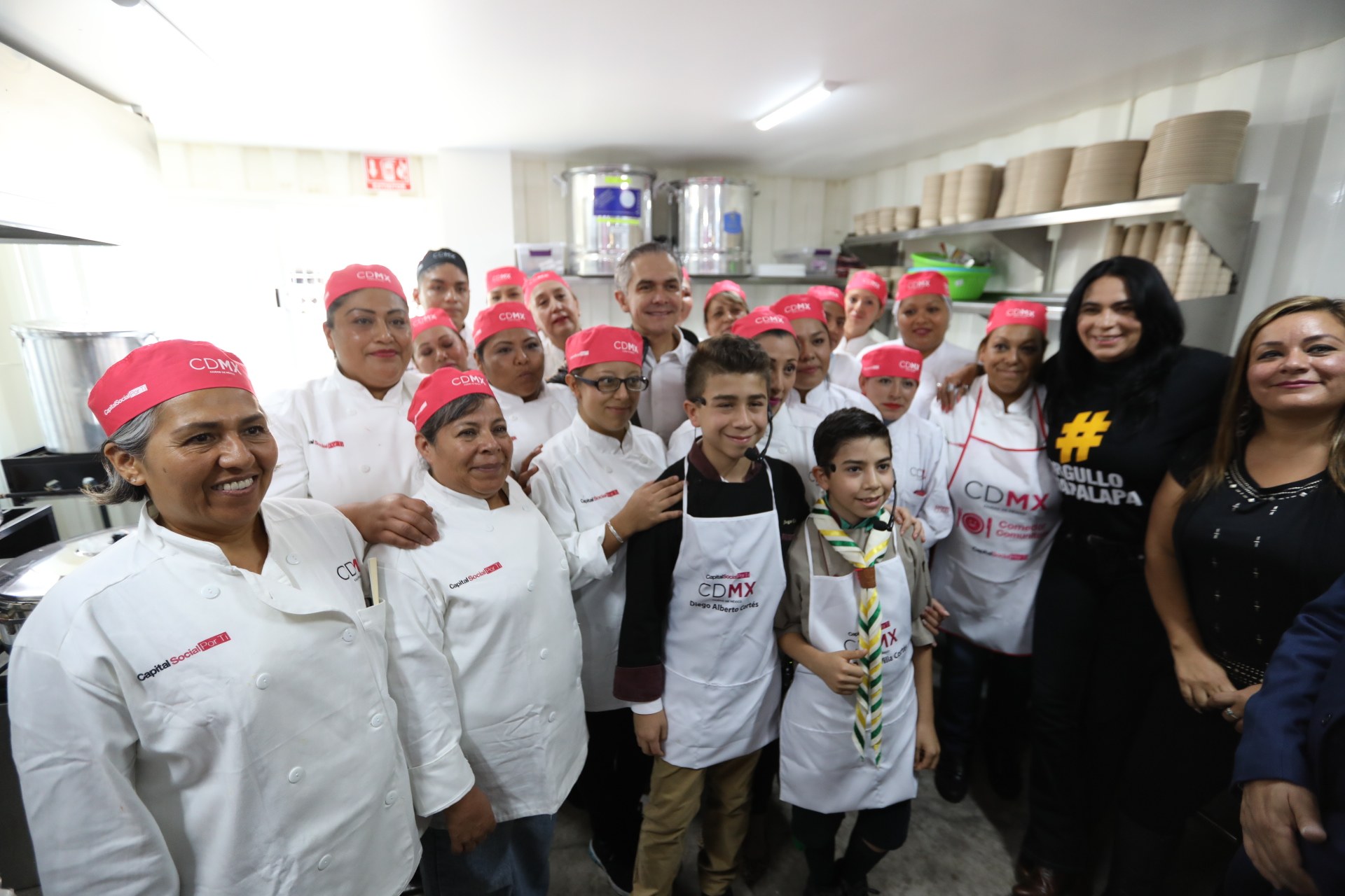 Eine Gruppe von Menschen in einer Küche, die fr├Âhlich in die Kamera l├Ąchelt und posiert, einige tragen K├Ąpchen, mit Utensilien links, Regalen mit Tellern und Sch├Ąlchen im Hintergrund und Deckenleuchten.