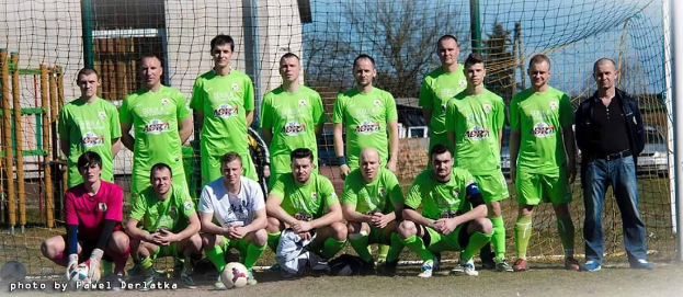 Gruppe von Männern in grünen T-Shirts auf einem Fußballfeld neben einem Torpfosten stehend, mit einem Ball auf dem Boden und Bäumen, Gebäuden und Fahrzeugen im Hintergrund.