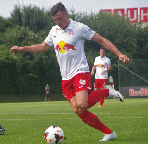Ein professioneller Fußballspieler kickt einen Ball auf einem Rasenfeld mit Bäumen und einem klaren blauen Himmel im Hintergrund, in der Nähe eines Schilds mit der Aufschrift "RB Leipzig."