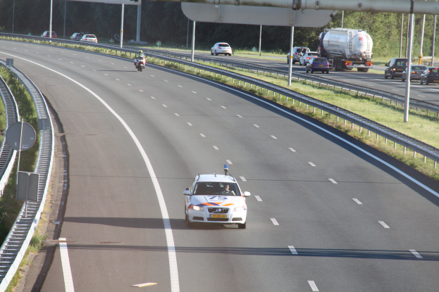 Ein Auto fährt auf einer Autobahn mit einem Polizeiwagen am Straßenrand, anderen Fahrzeugen, Leitplanken, Pfosten, Planken und Bäumen im Hintergrund.