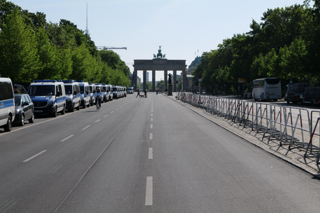 Eine Reihe von Polizeiwagen, die auf der Seite einer Straße vor dem Brandenburger Tor in Berlin, Deutschland, geparkt sind, mit Menschen, die Fahrräder fahren und in der Nähe stehen, Absperrungen, Bäumen und einem Bogen mit Statuen im Hintergrund.