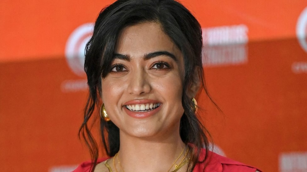 Frau in rotem Kleid mit elegantem Dutt und dezentem Make-up, die in die Kamera lächelt, vor einem gelben Banner mit der Aufschrift 'Sonam Kapoors beste Schönheits Looks vom Cannes Film Festival.'